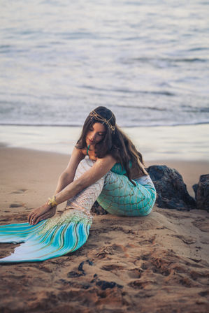 Person dressed as a mermaid in a teal scaled tail with flowing fin, wearing a gold headpiece and starfish bracelet, sitting on a sandy beach by gentle ocean waves at dusk