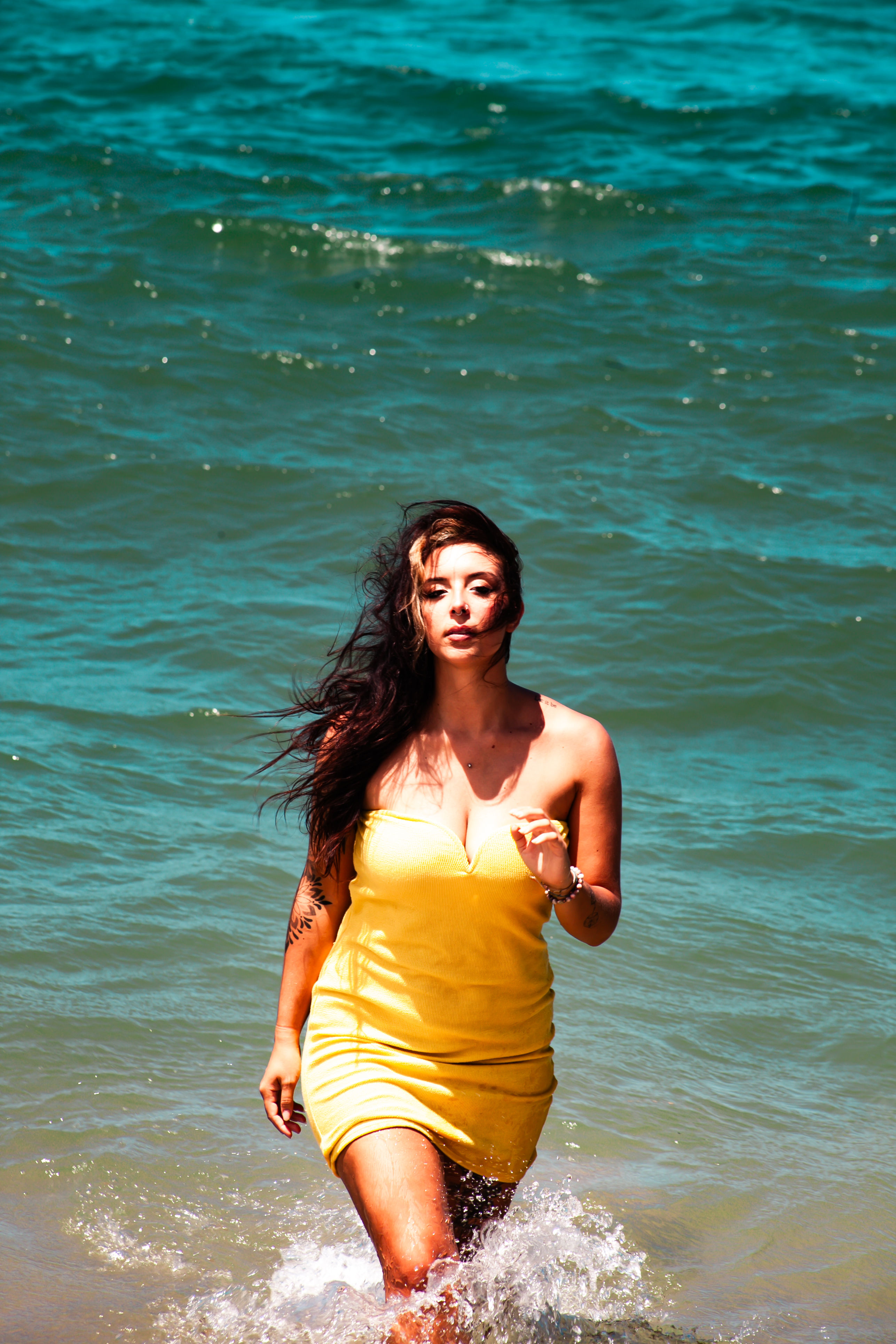 Woman in a yellow sundress walking out of turquoise ocean with splashing water and wind-blown hair, sunny beach vibe.