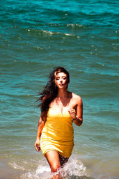 Woman in a yellow sundress walking out of turquoise ocean with splashing water and wind-blown hair, sunny beach vibe.