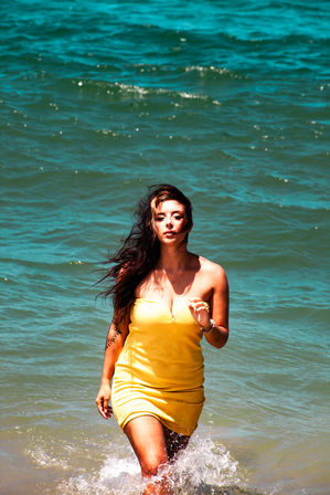 Woman in a yellow sundress walking out of turquoise ocean with splashing water and wind-blown hair, sunny beach vibe.