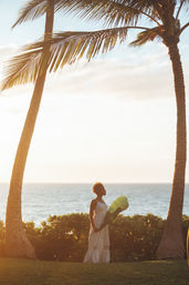 Silhouette of a woman in a white sundress holding a large tropical leaf between two palm trees on an oceanfront lawn at golden sunset.