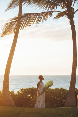 Silhouette of a woman in a white sundress holding a large tropical leaf between two palm trees on an oceanfront lawn at golden sunset.