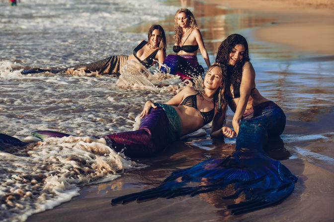 Four people wearing colorful mermaid tails lounging and smiling at the water's edge on a sunlit sandy beach as gentle waves wash around them.