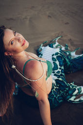 Smiling woman in a green mermaid tail and bikini top lounging on a sandy beach, adorned with layered pearl and shell jewelry, looking over her shoulder at the camera.