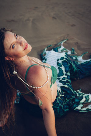 Smiling woman in a green mermaid tail and bikini top lounging on a sandy beach, adorned with layered pearl and shell jewelry, looking over her shoulder at the camera.
