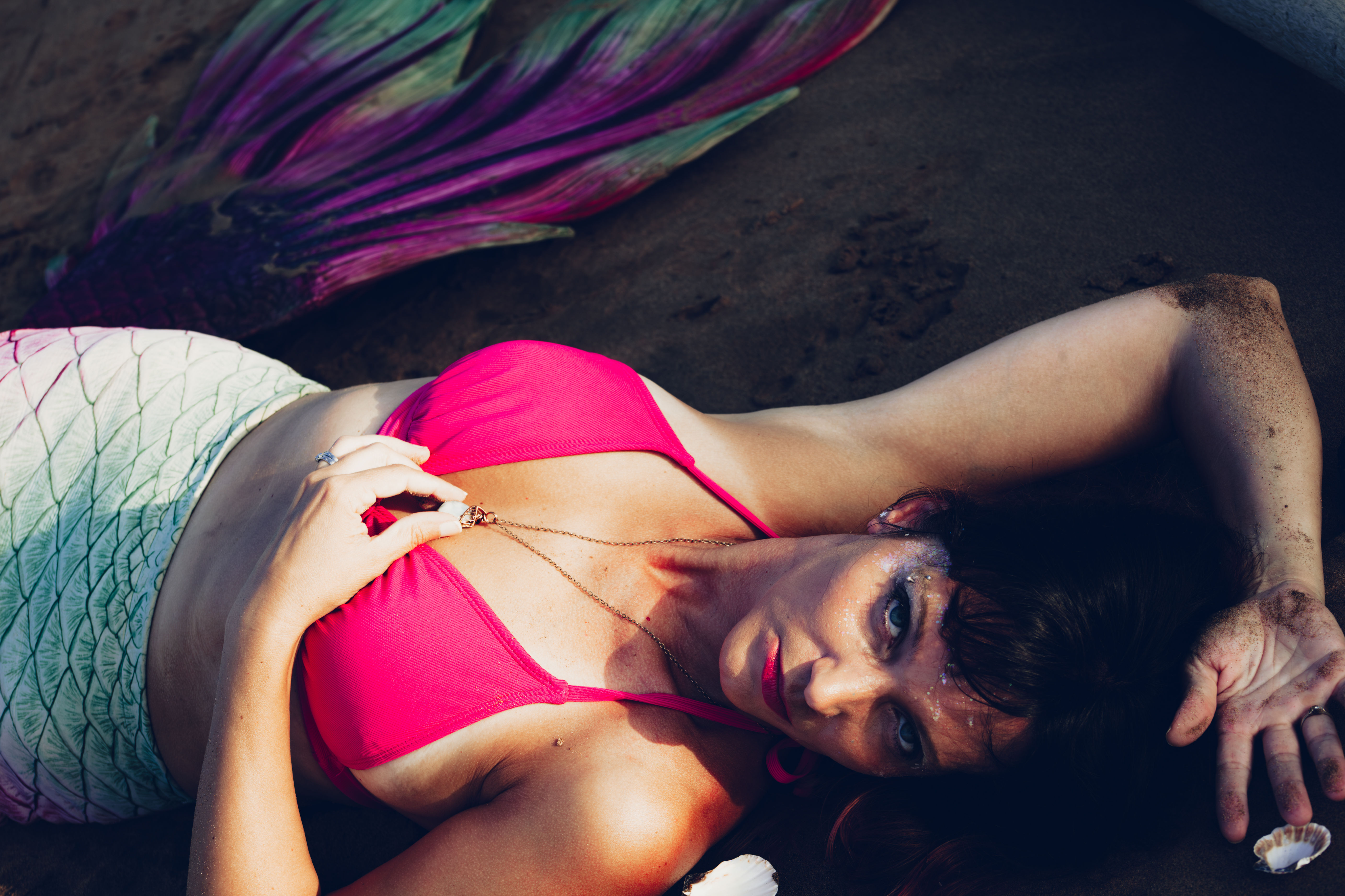 Person in mermaid costume reclining on dark sand with an iridescent green-pink tail, bright pink bikini top, holding a pendant necklace, glittered makeup, sandy arms and a nearby seashell.