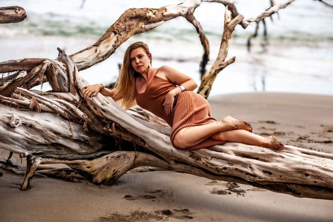 Woman in a rust-colored knit dress reclining barefoot on large driftwood on a sandy beach, wind-swept hair and relaxed coastal portrait — beach driftwood photoshoot