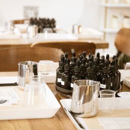 Scent-blending workshop table with dark glass dropper bottles, stainless steel jugs, clear glass beakers and white trays on a wooden table, ready for DIY perfume mixing.