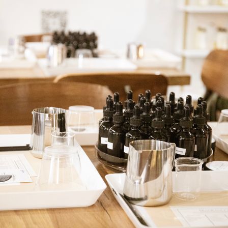 Scent-blending workshop table with dark glass dropper bottles, stainless steel jugs, clear glass beakers and white trays on a wooden table, ready for DIY perfume mixing.