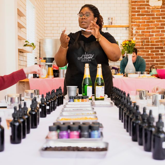 Instructor leading an artisan candle-making and scent-blending workshop in a cozy studio, standing behind a table lined with rows of small amber bottles, pigments, trays of ingredients and two sparkling beverage bottles against white subway tile and exposed brick.