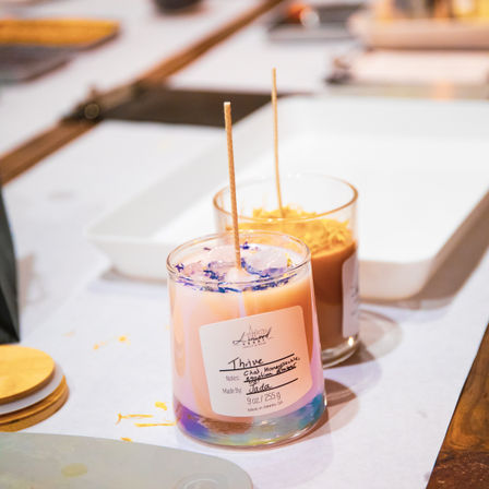 Two artisanal glass-jar candles with long wicks and dried flower petals—a pastel pink candle with a handwritten label in front and a tan candle behind—sitting on a white craft table at a candle-making workshop.