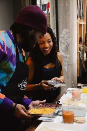 Two smiling participants in aprons at an indoor artisan candle-making workshop comparing trays of colorful additives—yellow wax shavings, dried flower petals and gray granules—over a worktable with candles and drinks.