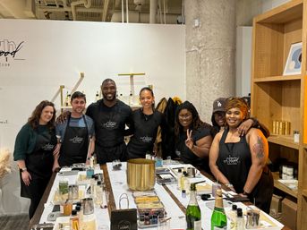 Smiling group of seven adults wearing black aprons at a candle-making workshop around a long table with jars, fragrance bottles, botanicals and champagne in a modern loft-style craft studio.