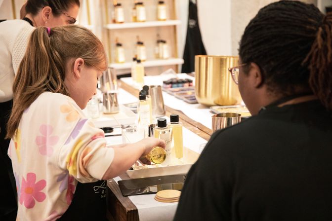 Young girl pouring golden liquid into a tray during a hands-on craft workshop, surrounded by bottles, metal tools, and gold-colored containers.