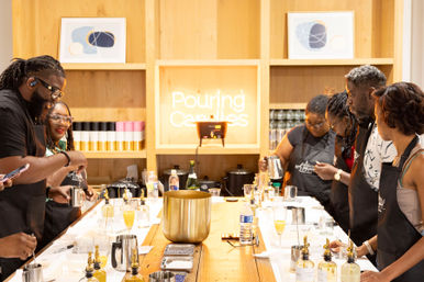 People in a candle-making workshop gathered around a long wooden table, pouring wax from pitchers into molds with fragrance bottles and tools, framed by a light-wood wall and a neon sign reading "Pouring Candles".