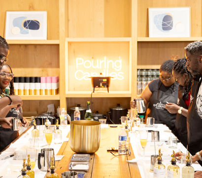 People in a candle-making workshop gathered around a long wooden table, pouring wax from pitchers into molds with fragrance bottles and tools, framed by a light-wood wall and a neon sign reading "Pouring Candles".