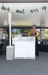 Smiling DJ in a black bikini with headphones standing behind a white booth under a covered backyard patio, speakers and a playful hanging banner for a poolside party.