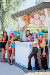 Six women in colorful wigs and swimwear pose by a DJ booth under a gazebo with a pink-and-cream balloon garland at a sunny backyard pool party, holding red cups.