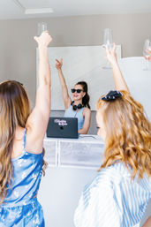 Female DJ in sunglasses and headphones playing from a laptop at a bright modern indoor party while friends raise wine glasses in a celebratory toast