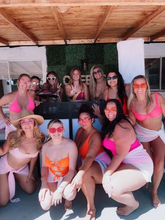 Group of women in bright swimsuits and playful sunglasses smiling and posing at a sunny backyard poolside patio under a wooden pergola with a green hedge backdrop