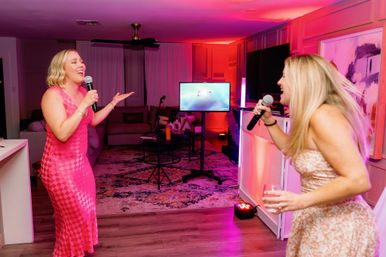 Two women singing karaoke in a living room with pink and purple party lights, microphones and a TV screen, one holding a wine glass — lively at-home karaoke night.