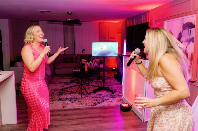 Two women singing karaoke in a living room with pink and purple party lights, microphones and a TV screen, one holding a wine glass — lively at-home karaoke night.