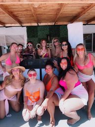 Group of women in bright swimsuits and playful sunglasses smiling and posing at a sunny backyard poolside patio under a wooden pergola with a green hedge backdrop