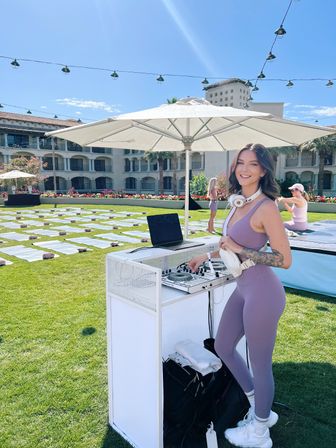 Smiling DJ in lavender activewear wearing headphones at a sunny outdoor resort lawn event, standing by a white DJ booth under a large umbrella with rows of yoga mats on the grass and string lights overhead.