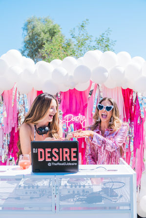 Outdoor summer party scene with two friends at a pink-themed DJ booth, white balloon cloud overhead, neon party sign, pink streamers and playful sunglasses.