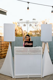 Outdoor patio DJ at a white booth during golden hour, wearing headphones and raising one hand, laptop with 'Flawlyss' sticker, covered speaker towers and string lights creating a backyard party vibe