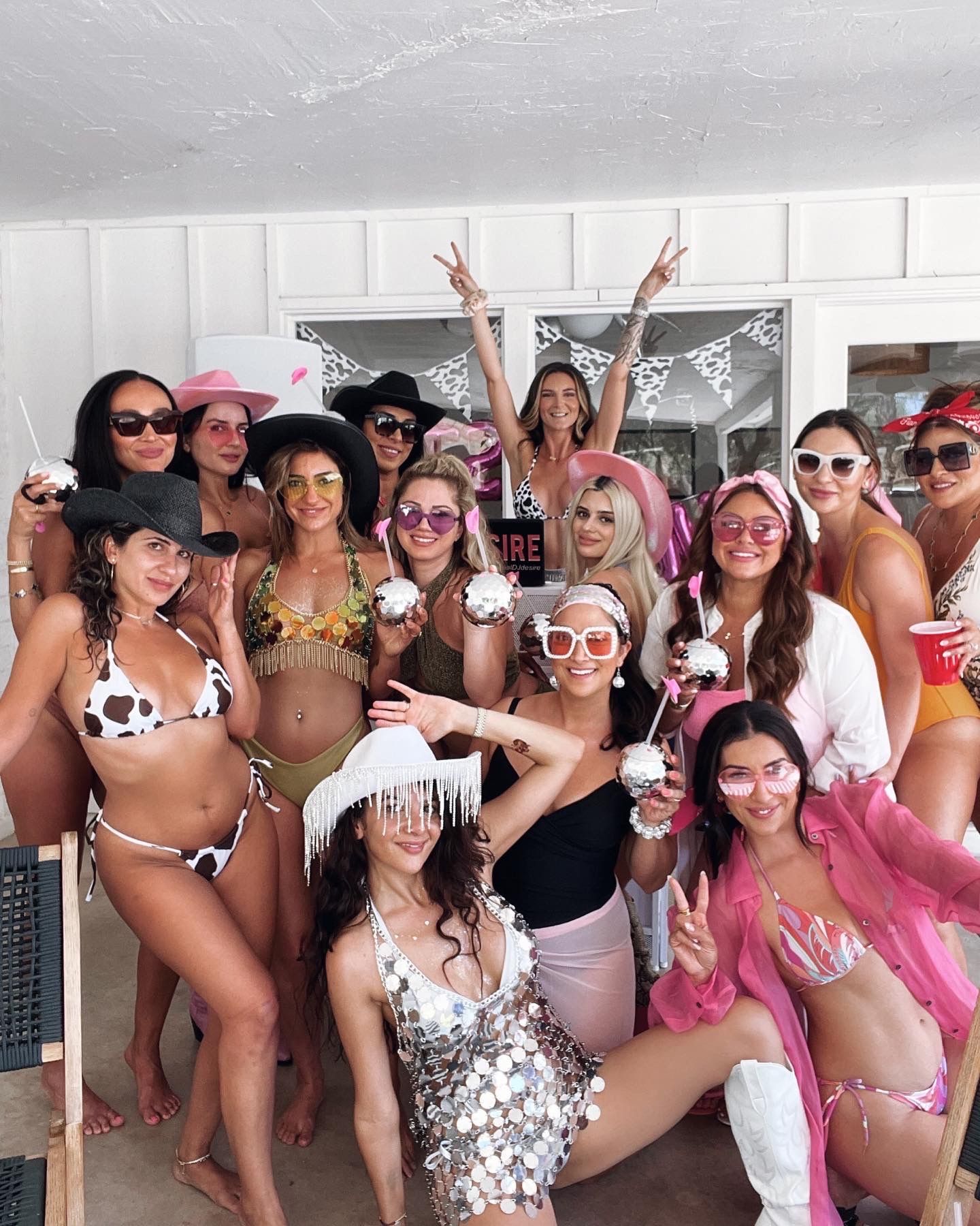 Cheerful group of women at a sunny poolside patio party in colorful swimsuits, pink hats and cowboy hats, holding disco-ball drinks and posing for a bachelorette-style celebration.