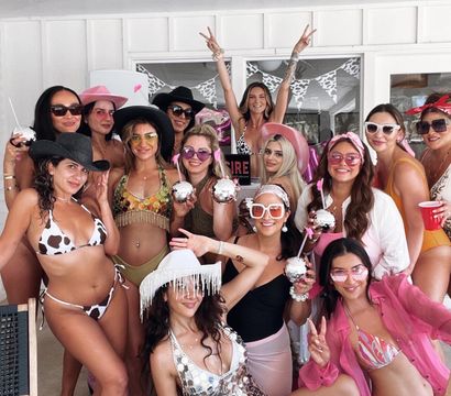 Cheerful group of women at a sunny poolside patio party in colorful swimsuits, pink hats and cowboy hats, holding disco-ball drinks and posing for a bachelorette-style celebration.