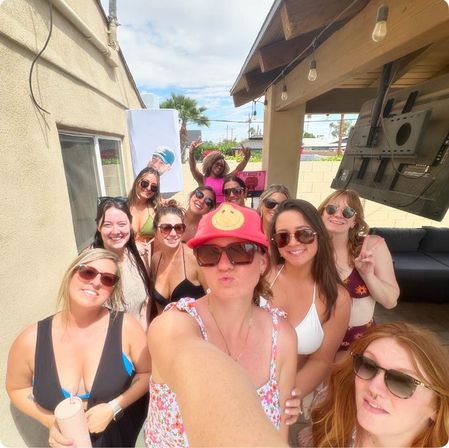 Selfie of a group of women in swimsuits and sunglasses at a sunny backyard patio party with string lights, palm trees, and a red smiley hat