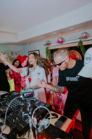 Energetic living-room party with a DJ controller in the foreground, a woman dancing with headphones around her neck and a man in sunglasses mixing music, disco balls and pink heart balloons overhead.