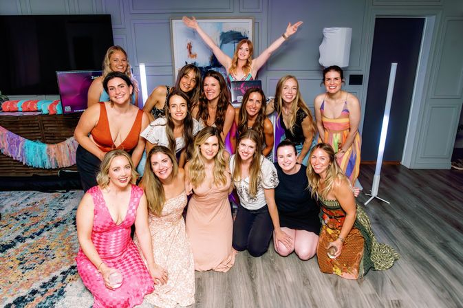 Smiling group of women posing for an indoor living-room celebration photo, wearing colorful dresses with festive tassel decor and drinks — joyful group shot.