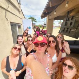 Selfie of a group of women in swimsuits and sunglasses at a sunny backyard patio party with string lights, palm trees, and a red smiley hat