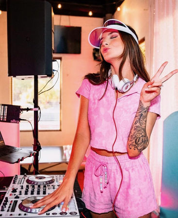 DJ in pink crop top and shorts wearing a visor and headphones, flashing a peace sign with a tattooed arm while mixing on a DJ controller at an indoor pastel-themed party.