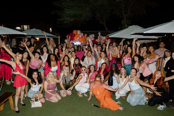 Large diverse group photo at a nighttime outdoor patio party, people in colorful pink and orange outfits cheering and waving in front of a balloon arch and patio umbrellas on a green lawn.