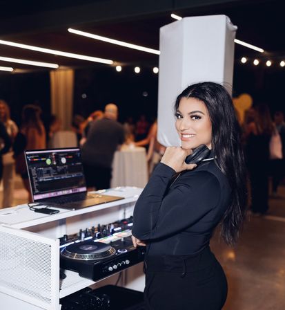 Smiling DJ at a white booth holding headphones over one shoulder, with a laptop and turntable controller, playing music at an indoor evening party under string lights with a blurred crowd in the background.