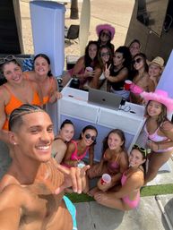 Smiling shirtless man takes a selfie with a lively group of friends in colorful bikinis at a sunny beachside party around a DJ booth with a laptop and speakers, red cups and pink cowboy hats visible.