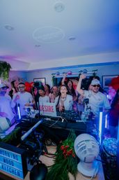 Energetic indoor party with a DJ at a packed booth, cheering guests, disco balls, balloons, colorful LED lighting and streaming cameras.