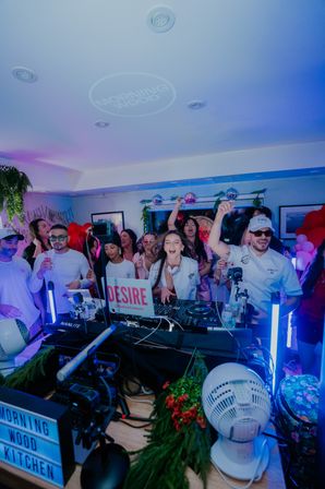 Energetic indoor party with a DJ at a packed booth, cheering guests, disco balls, balloons, colorful LED lighting and streaming cameras.