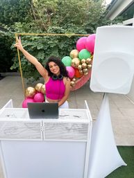 Smiling DJ with headphones raising a peace sign behind a white DJ booth with laptop and speaker on a tiled patio, colorful pink, green and gold balloon arch and leafy garden backdrop.