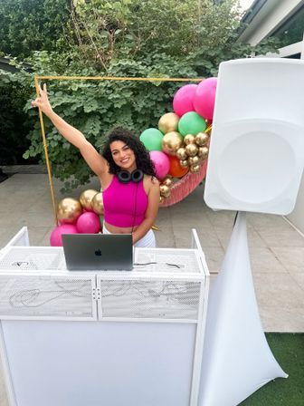 Smiling DJ with headphones raising a peace sign behind a white DJ booth with laptop and speaker on a tiled patio, colorful pink, green and gold balloon arch and leafy garden backdrop.