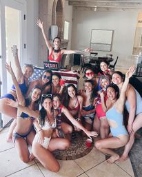 Group of friends in colorful swimsuits cheering and posing at an indoor patriotic pool-party gathering with a DJ booth draped in an American flag, red solo cups, sunglasses and red-white-blue accessories in a bright vacation home living area.