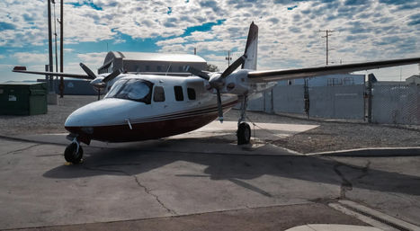 Small twin-engine propeller aircraft parked on a tarmac at a small airport, tied down near hangars and a chain-link fence under a dramatic cloud-streaked blue sky.