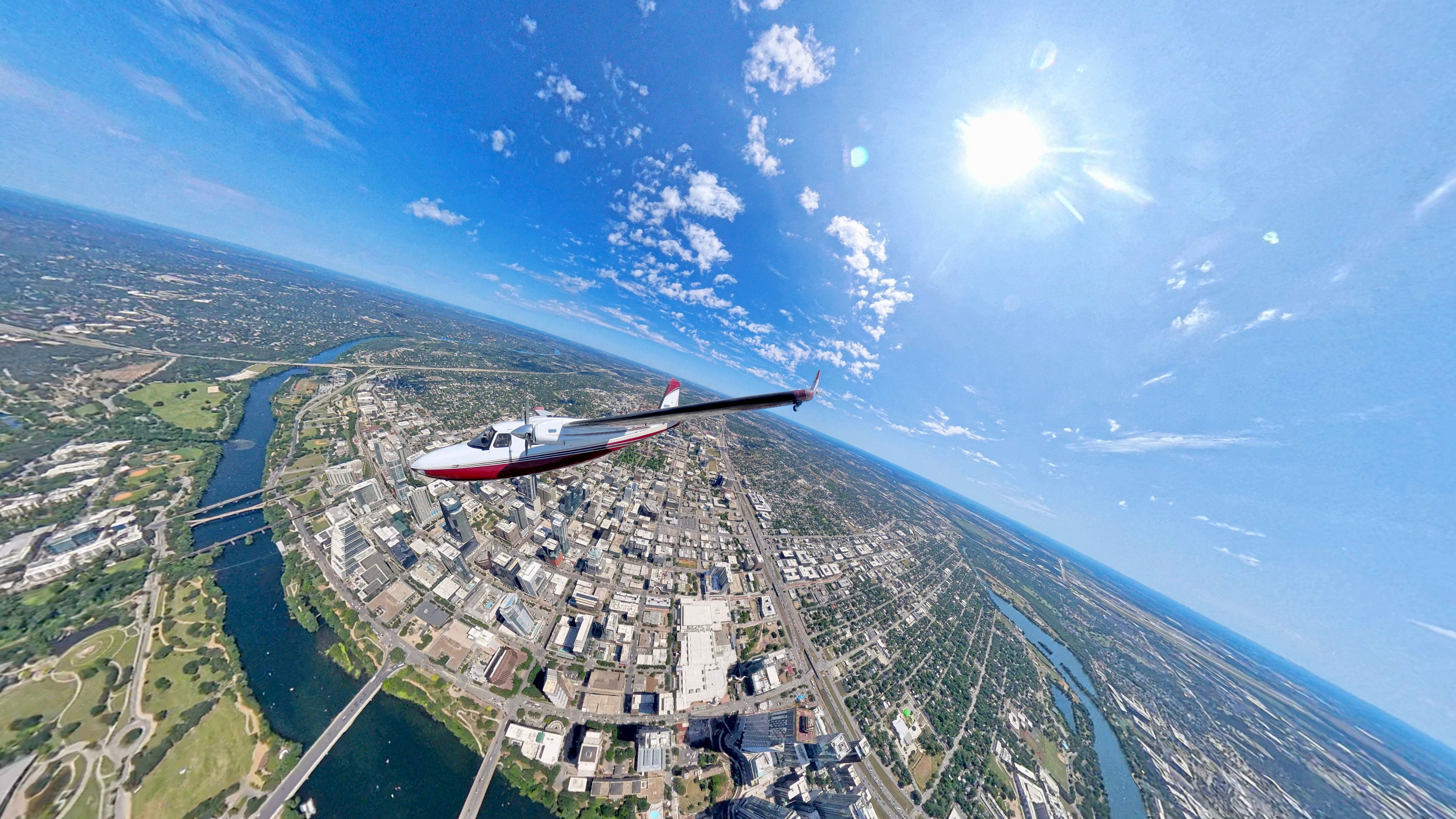 Aerial view of a small red-and-white plane soaring over a sunlit downtown, winding river and bridges under a bright blue sky with scattered clouds.