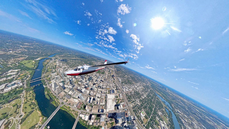 Aerial view of a small red-and-white plane soaring over a sunlit downtown, winding river and bridges under a bright blue sky with scattered clouds.