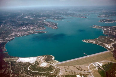 Aerial view of a large turquoise reservoir held by an earthen dam, with curving peninsulas, winding shoreline roads and scattered suburban development under a hazy sky.