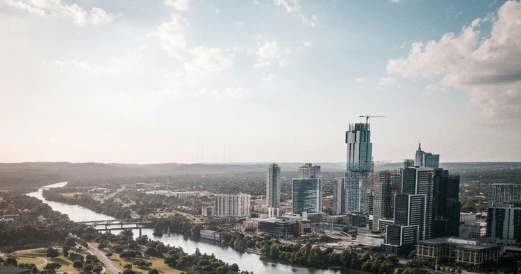 Aerial downtown riverfront skyline with modern high-rise towers, a winding river and bridges flanked by green parks under a bright, partly cloudy sky.